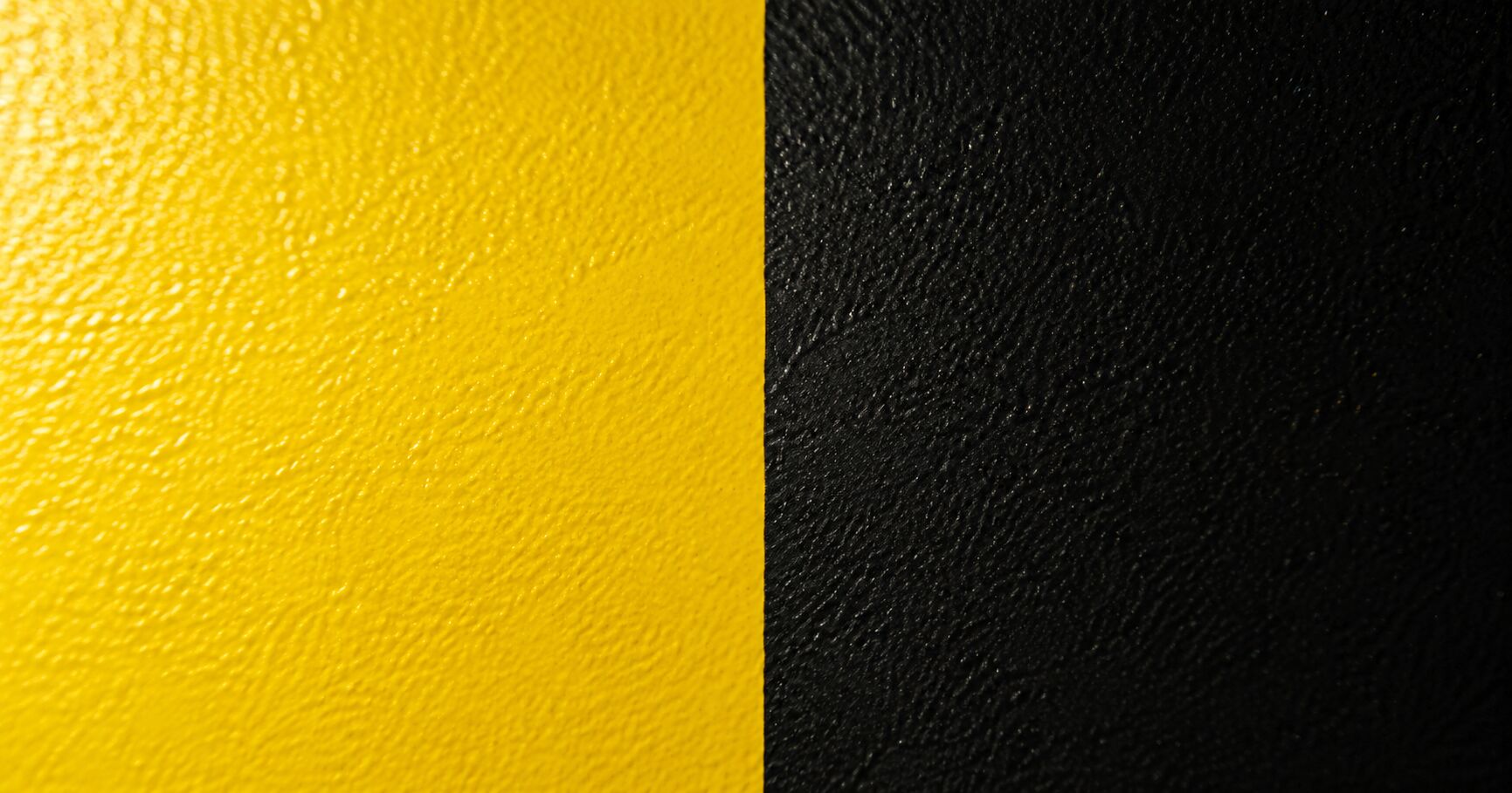 A macro of cured powder coated metal split vertically, half saturated industrial yellow, half deep ink black, fine matt orange peel texture